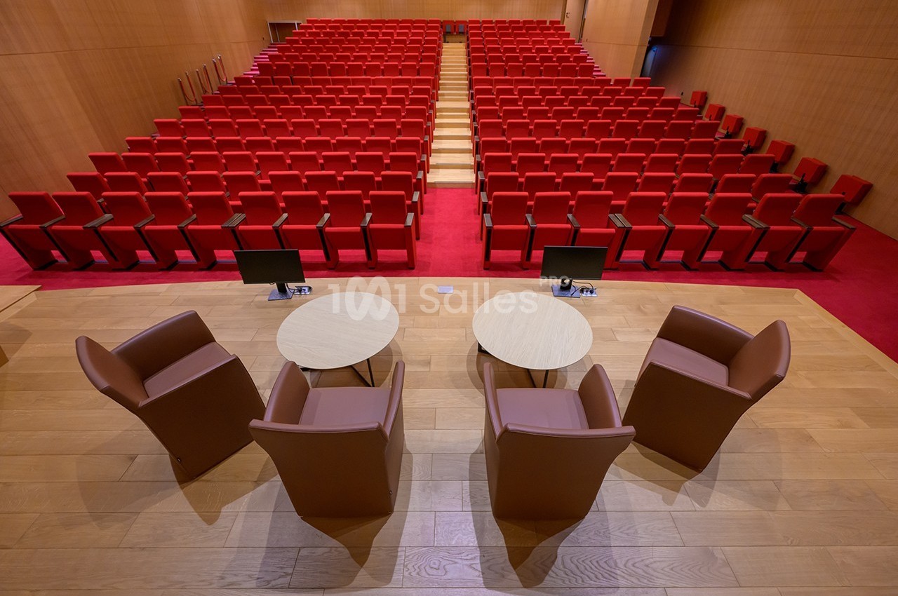 Salle de conférence avec rangées de sièges rouges, vue depuis une scène équipée de fauteuils et tables rondes.
