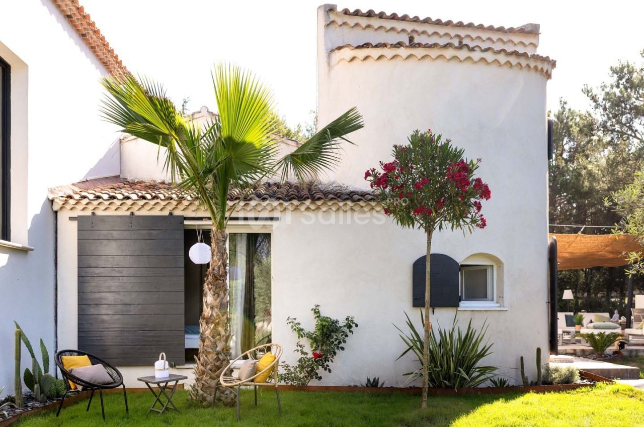 Façade d'une maison méditerranéenne avec jardin, palmiers, chaises et table sur une pelouse.