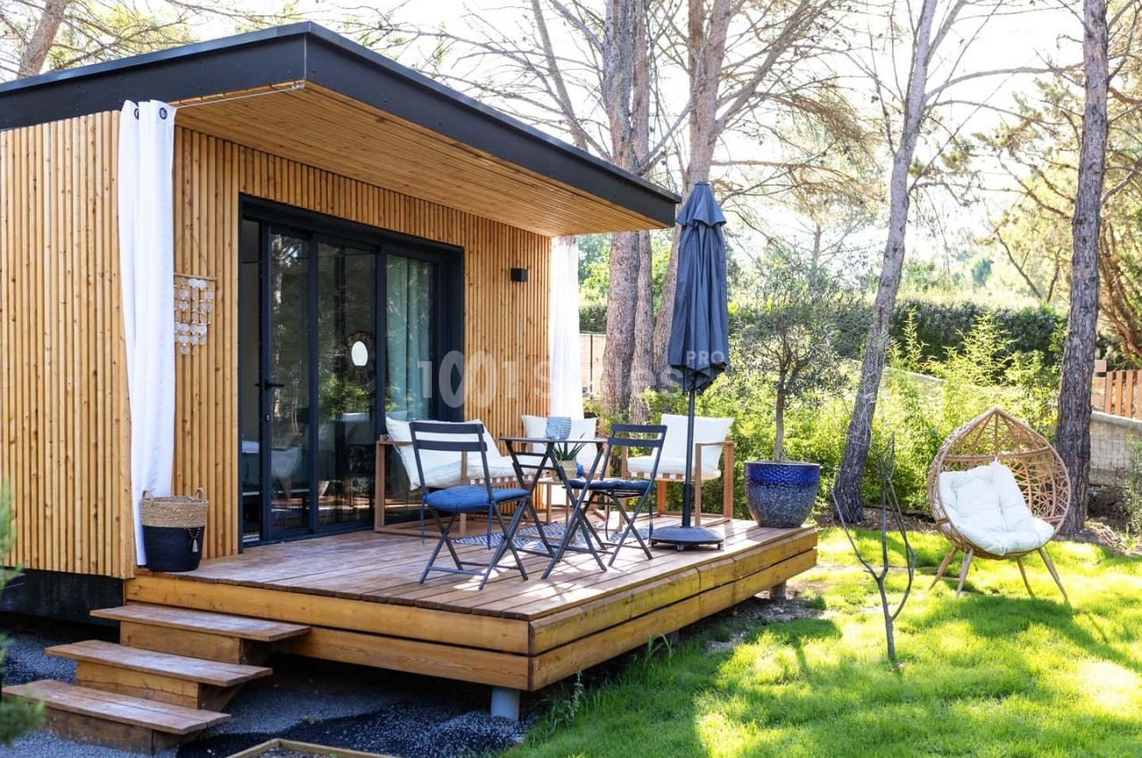 Terrasse en bois avec mobilier de jardin devant une petite maison moderne entourée de verdure et d'arbres.