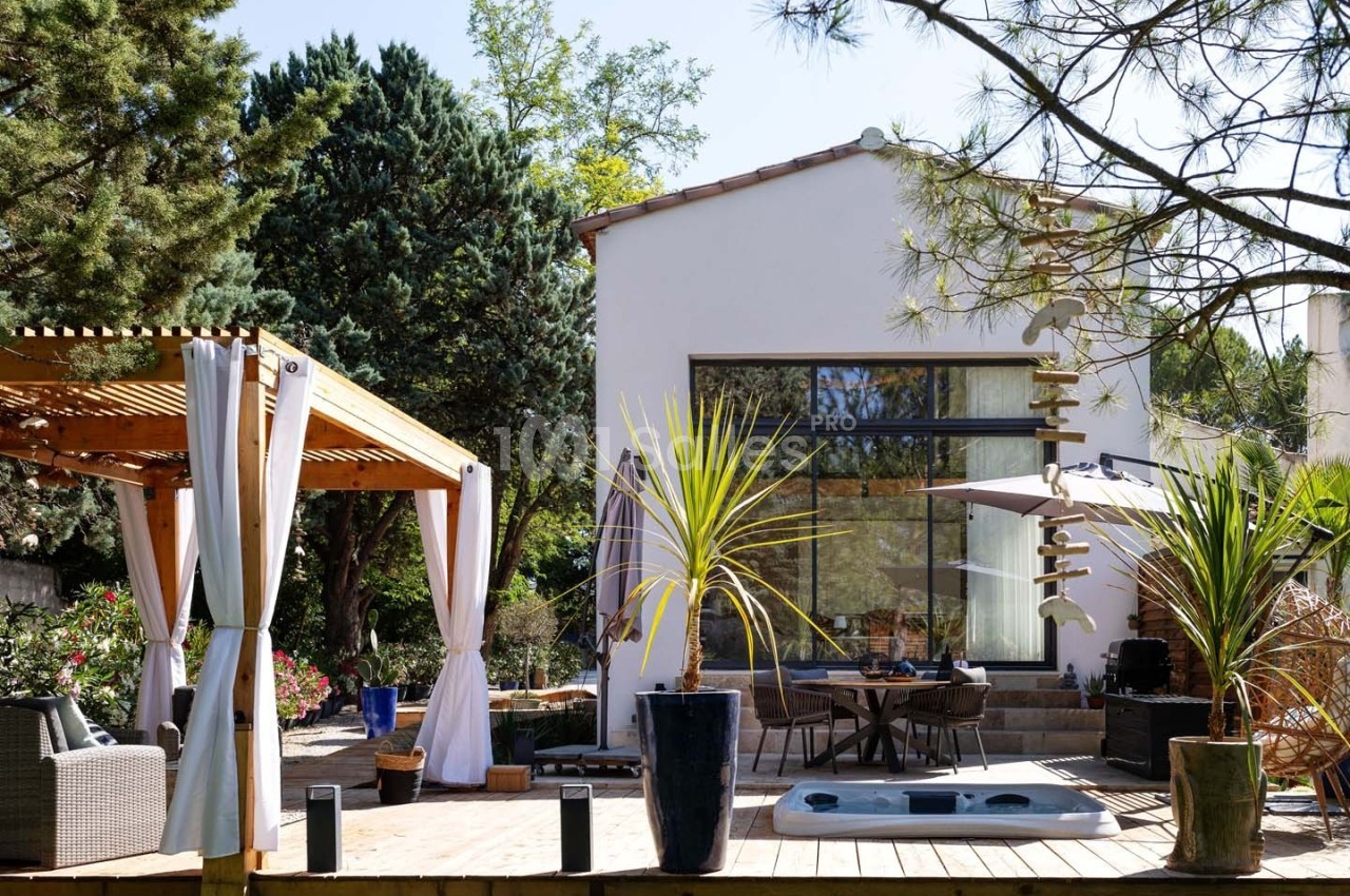 Maison moderne avec terrasse en bois, pergola, jacuzzi et végétation environnante sous un ciel dégagé.