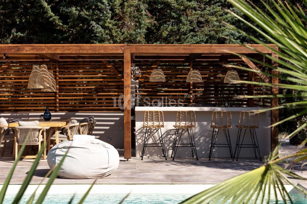 Espace extérieur avec pergola en bois, bar avec tabourets, table à manger, pouf blanc et piscine au premier plan.