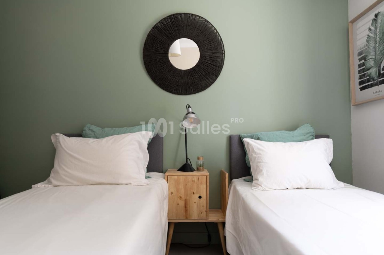 Deux lits simples avec des draps blancs, une table de chevet en bois, une lampe et un miroir rond sur un mur vert.