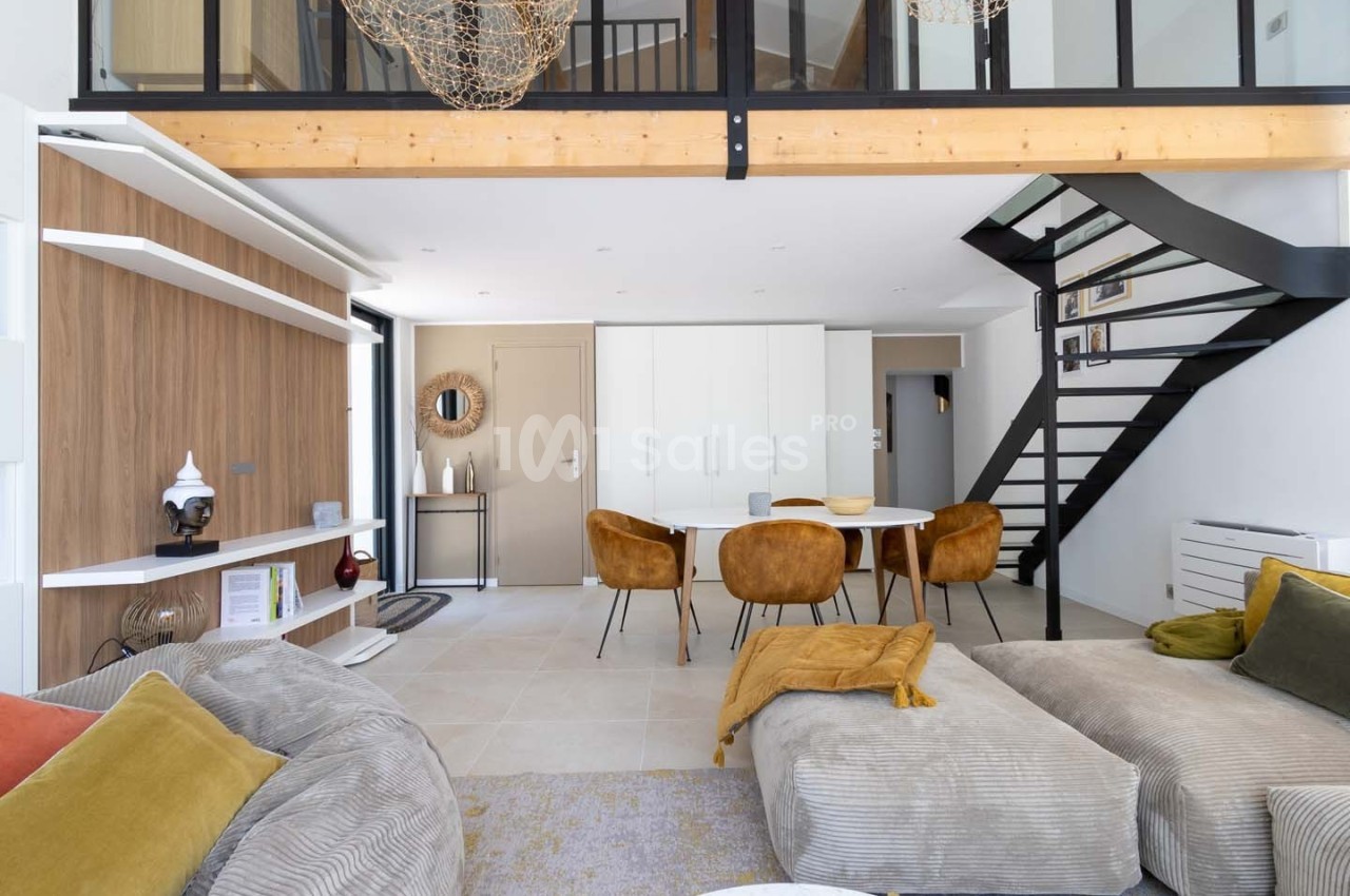 Salon moderne avec mezzanine, canapé beige, table à manger entourée de chaises moutarde et escalier en métal noir.