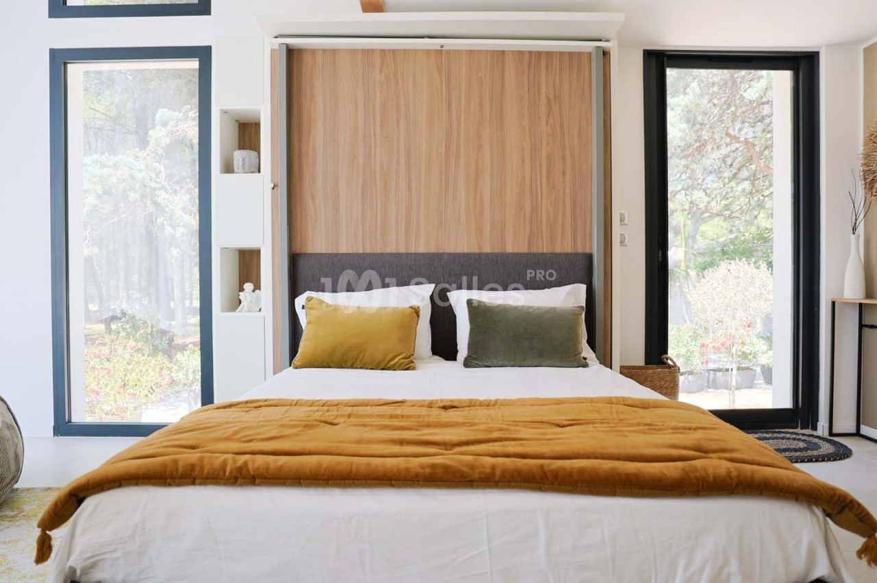 Lit double avec une couverture ocre dans une chambre lumineuse, dotée de grandes fenêtres et d'un décor minimaliste.