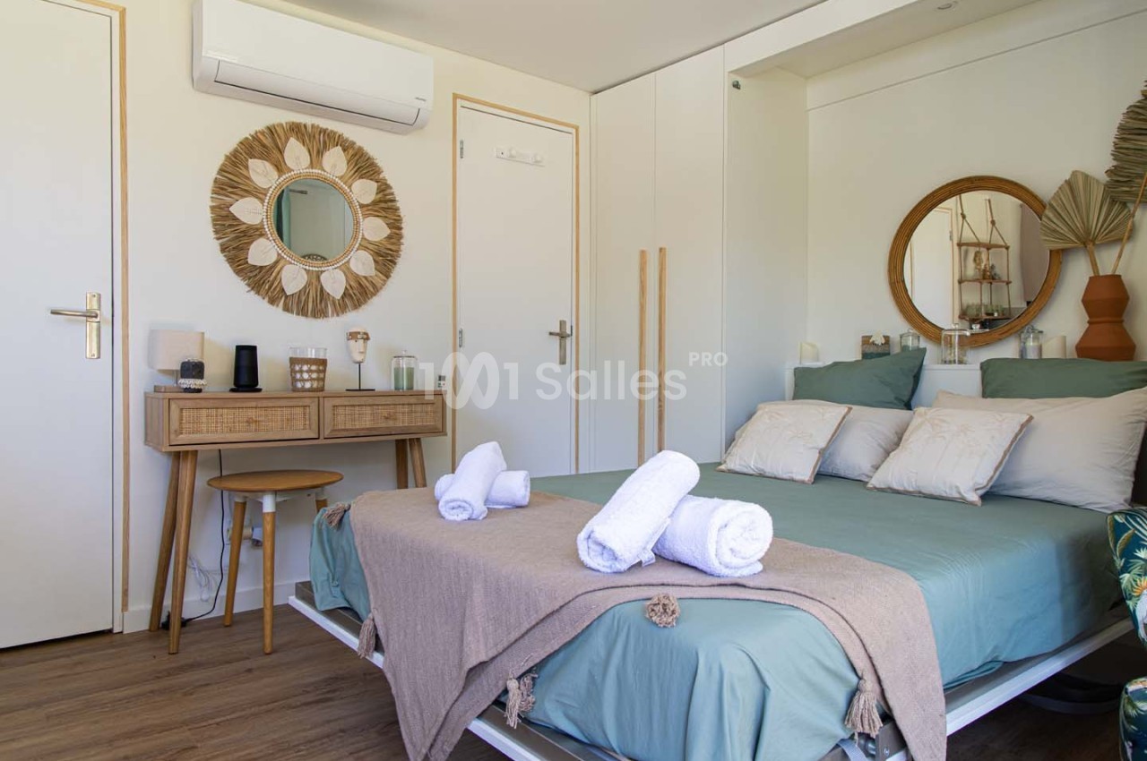 Chambre lumineuse avec lit double, linge plié, bureau en bois, miroir décoratif et climatisation murale.