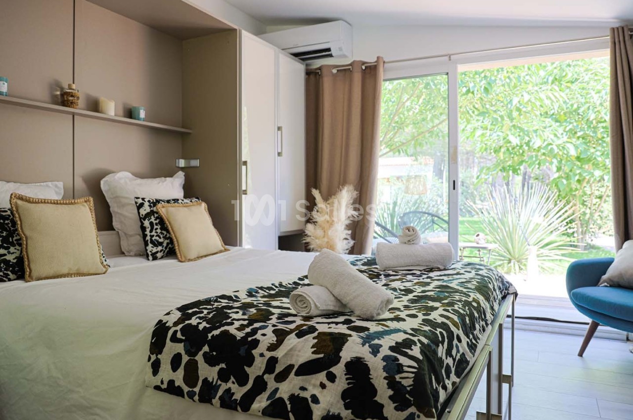 Chambre lumineuse avec lit double, coussins décoratifs, serviettes pliées et grande baie vitrée donnant sur un jardin.