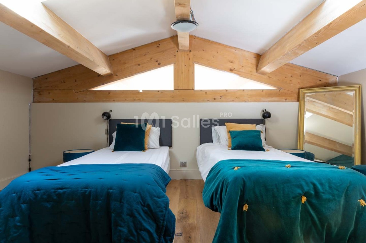 Deux lits simples avec couvertures bleues dans une chambre mansardée aux poutres apparentes et miroir au sol.