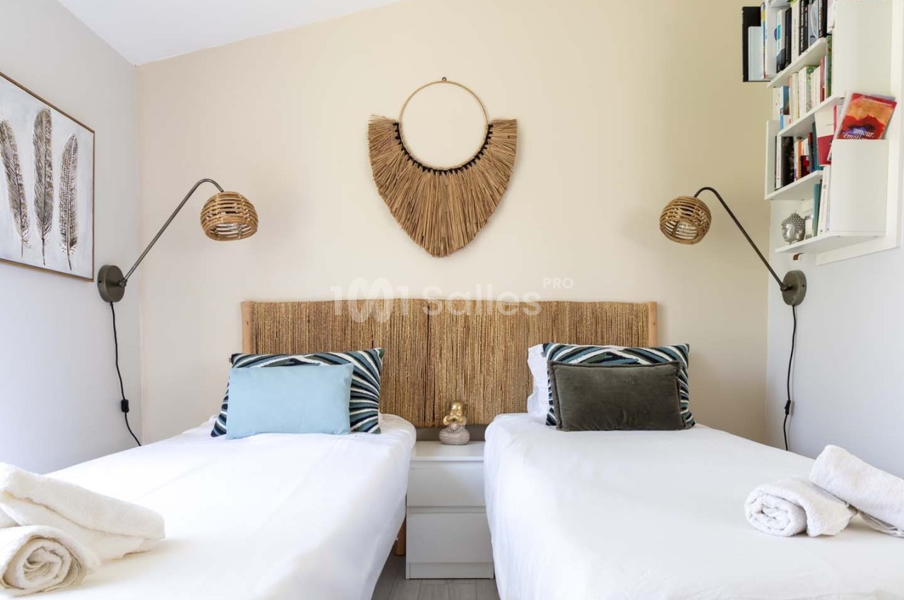 Deux lits simples avec linge blanc, coussins décoratifs, lampes murales et décoration murale en fibres naturelles.