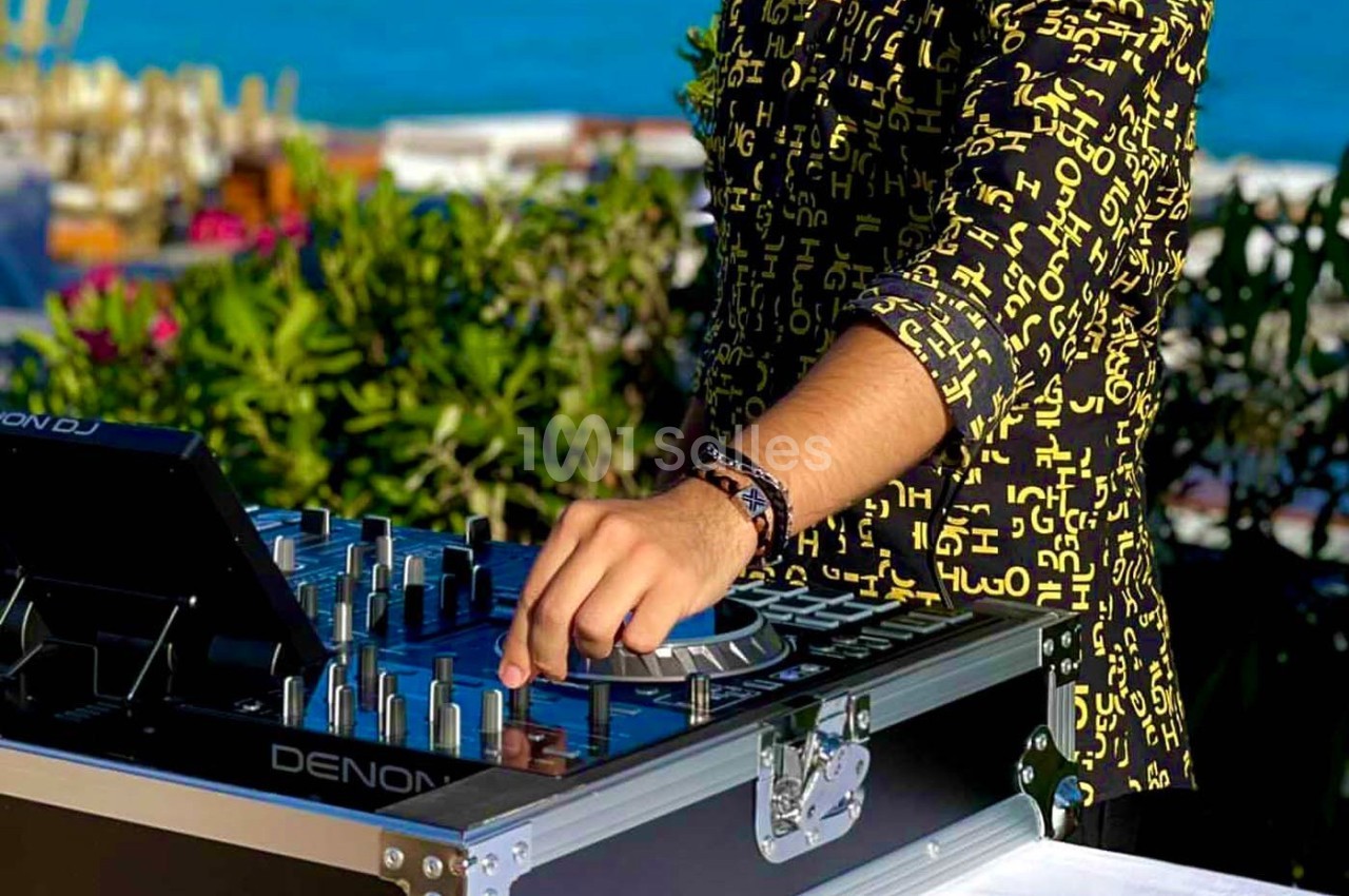 Un DJ ajuste une table de mixage en plein air, entouré de verdure et avec une vue sur la mer en arrière-plan.