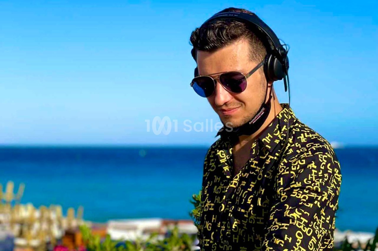 Un DJ portant un casque audio et des lunettes de soleil mixe en extérieur avec la mer en arrière-plan.