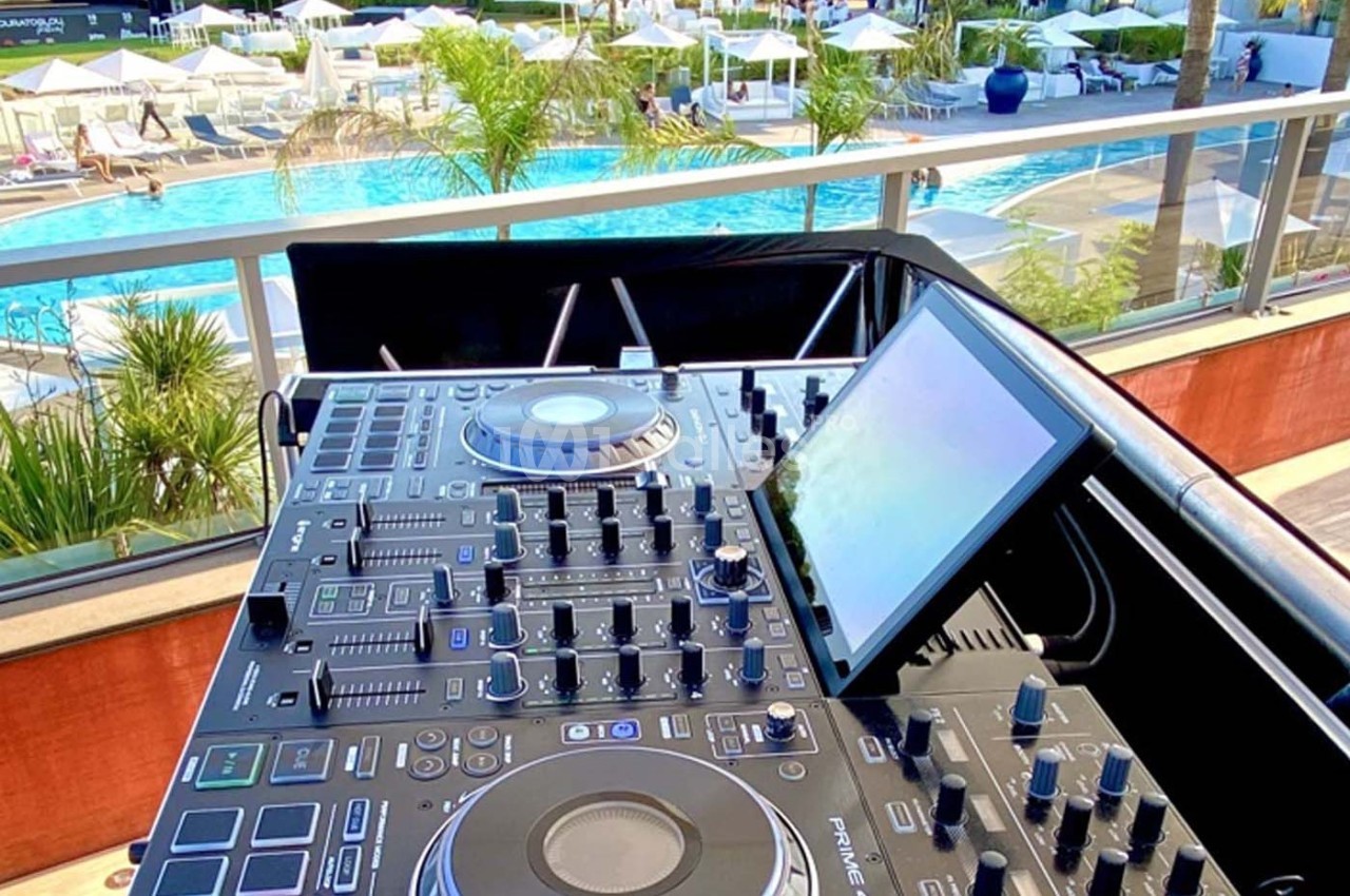 Table de mixage DJ installée en extérieur, avec vue sur une piscine entourée de transats et de parasols.