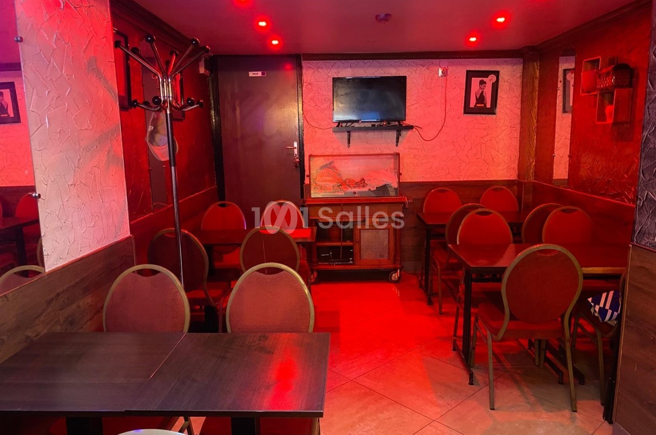 Salle de restaurant avec des tables et chaises alignées, éclairée par une lumière rouge et une télévision fixée au mur.