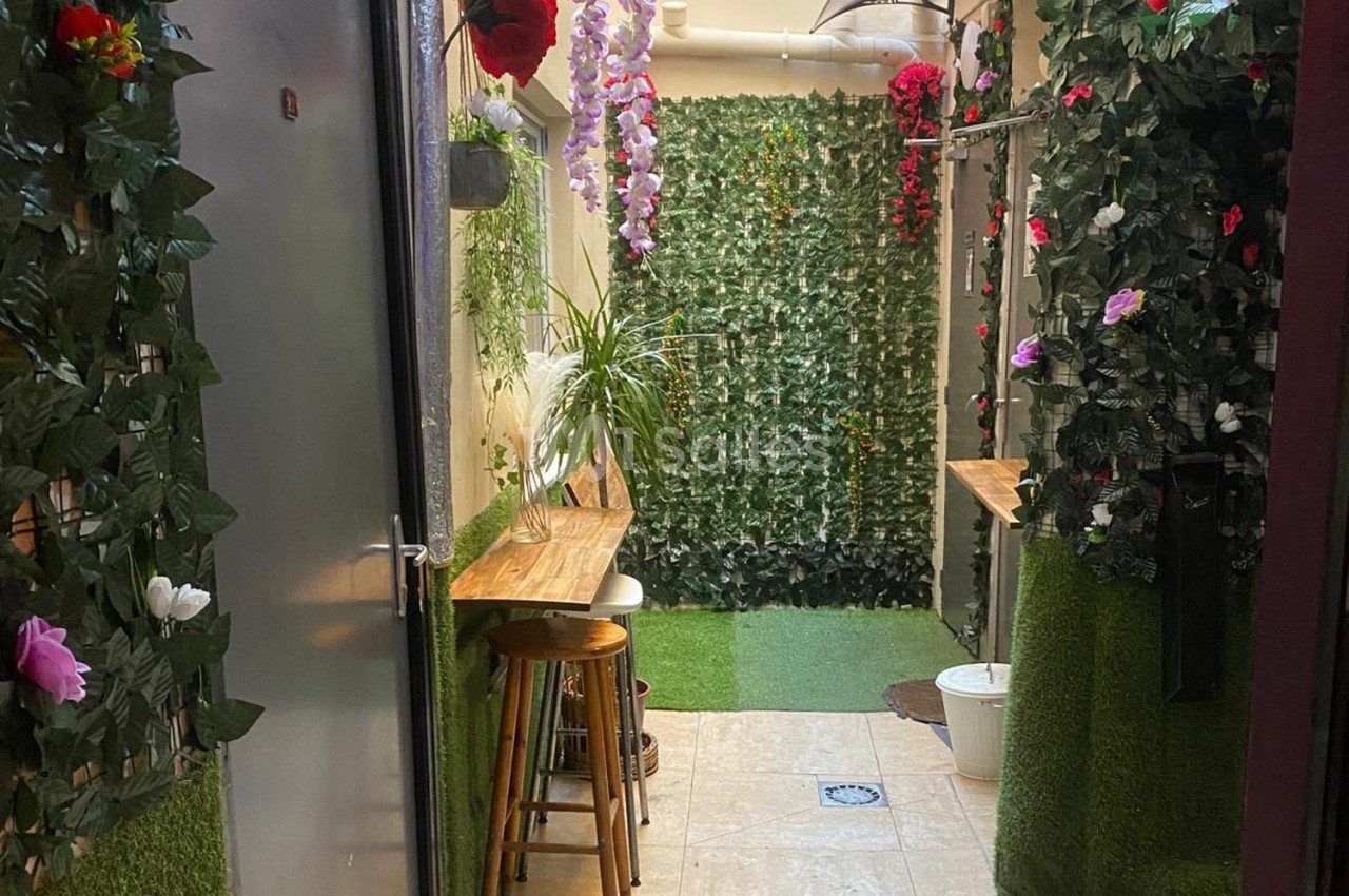 Couloir étroit décoré de plantes artificielles, fleurs colorées et comptoirs en bois avec tabourets.