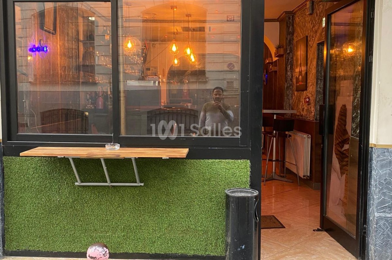Façade d'un café avec une table extérieure en bois, revêtement en gazon synthétique et intérieur éclairé visible.