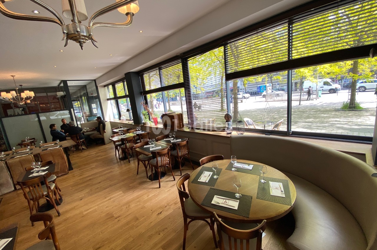 Salle de restaurant lumineuse avec grandes baies vitrées, tables en bois et banquettes arrondies près des fenêtres.