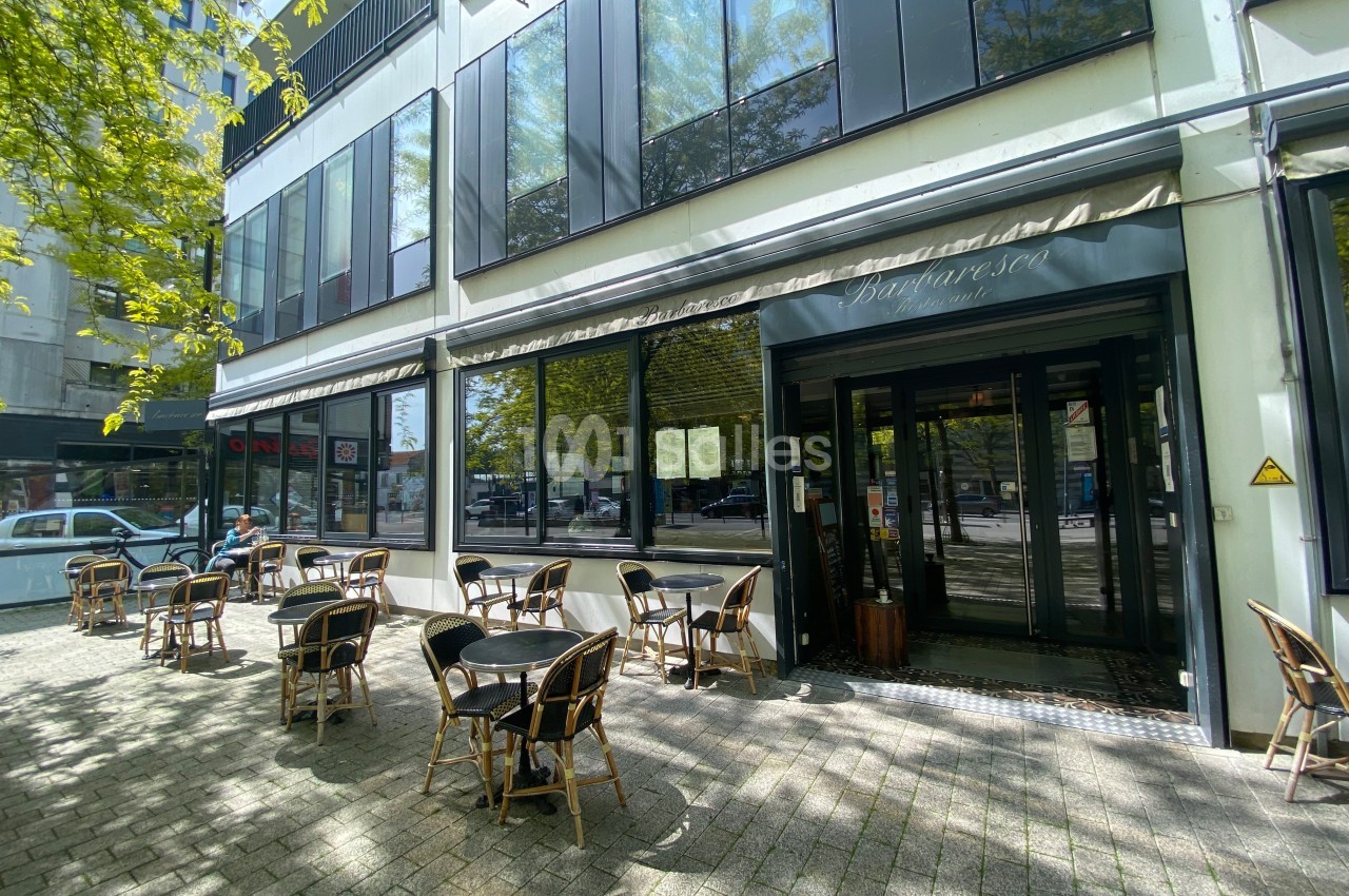 Terrasse d'un café avec des tables et chaises en extérieur, située devant une façade vitrée sous un auvent.