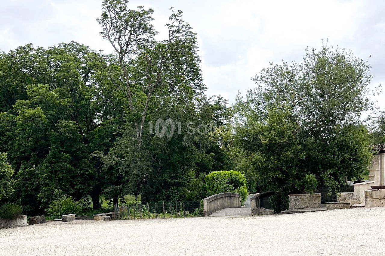 Place ouverte avec sol en gravier, entourée d'arbres verdoyants et de petits murets en pierre.