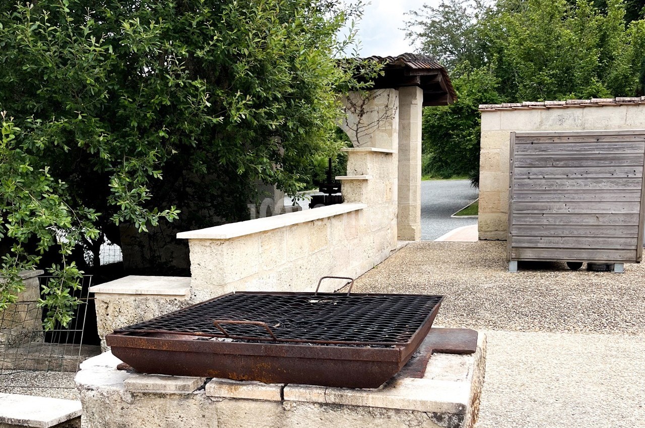 Barbecue en métal rouillé installé sur un muret en pierre, entouré d'arbres et de structures extérieures.