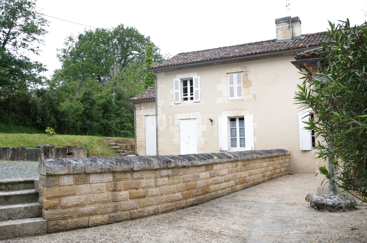 Maison en pierre beige avec volets blancs, entourée d'un muret en pierre et d'une végétation verdoyante.