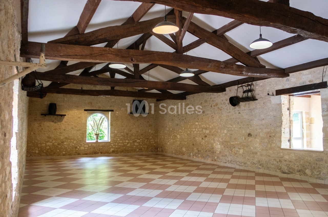 Salle vide avec murs en pierre, poutres en bois apparentes et carrelage beige et rose, éclairée par des fenêtres.