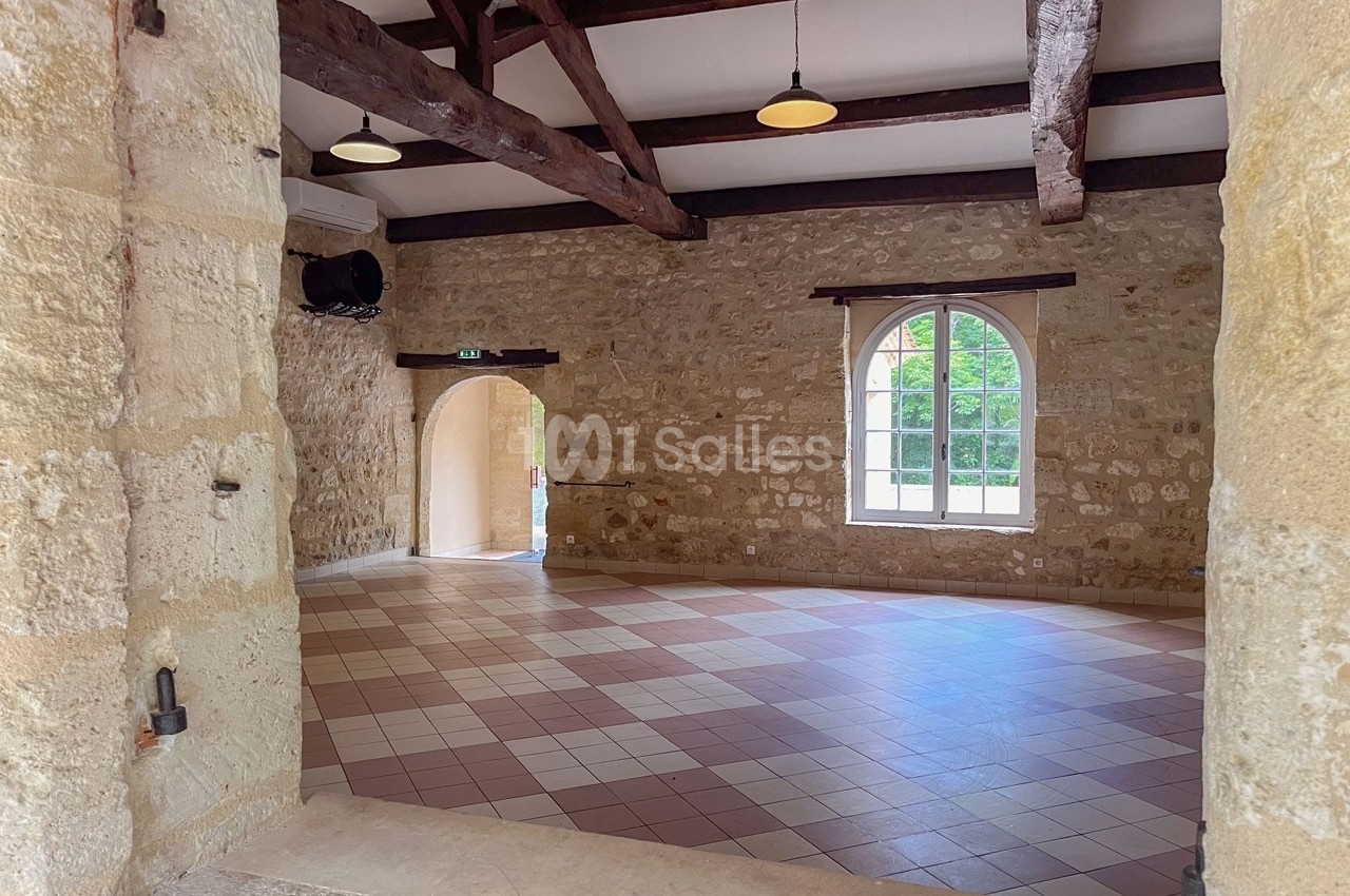 Salle en pierre avec poutres apparentes, carrelage au sol, grande fenêtre et porte voûtée ouverte sur l'extérieur.