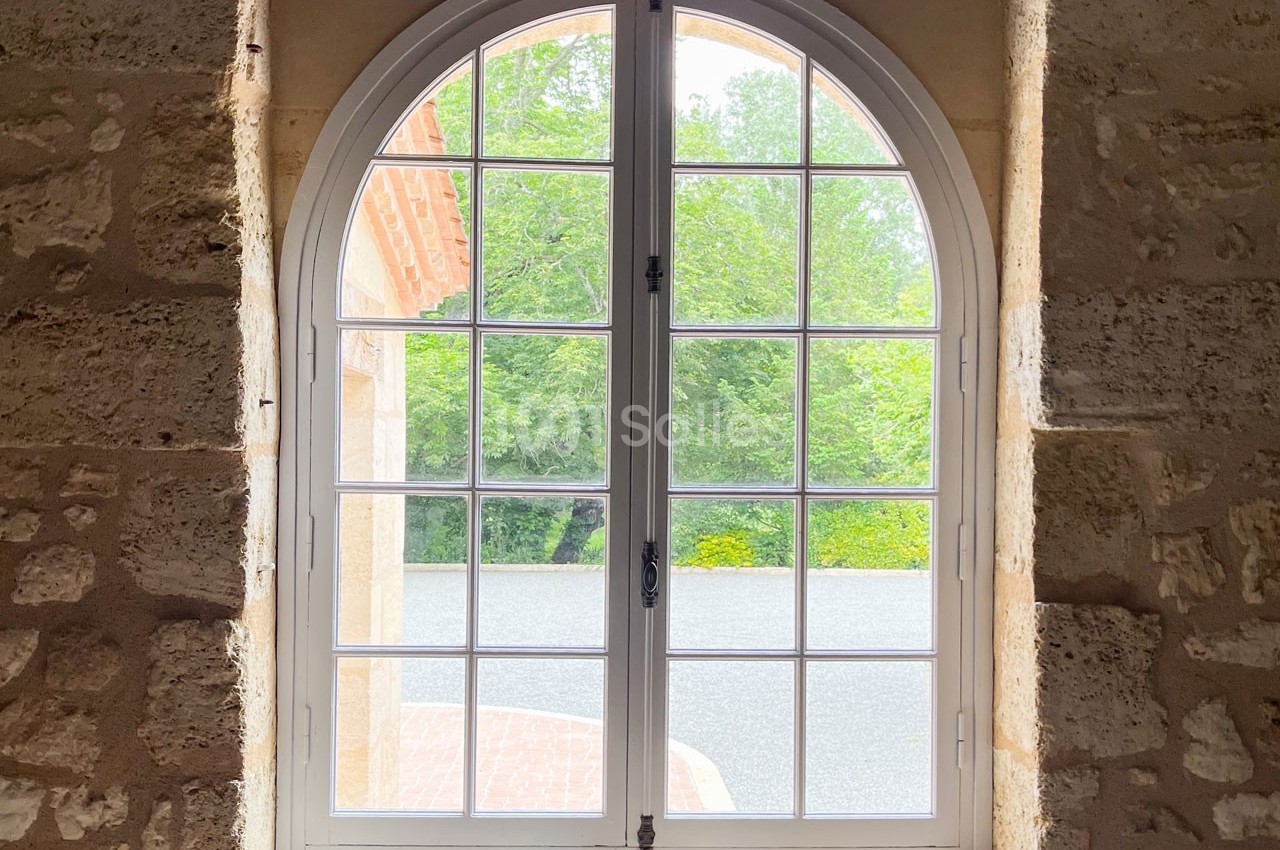 Fenêtre cintrée en bois blanc donnant sur un jardin verdoyant, encadrée par un mur en pierre beige.