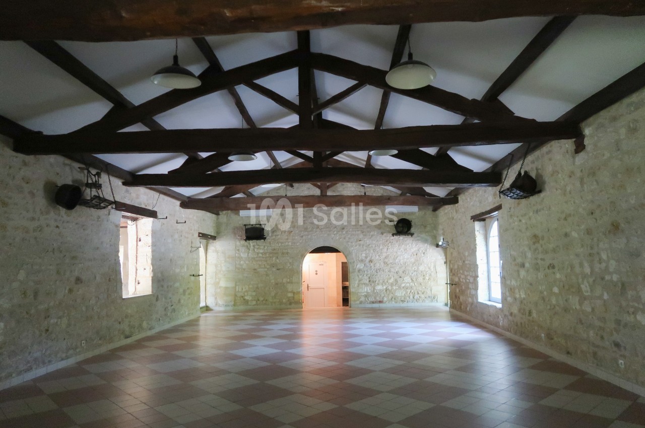Salle intérieure en pierre avec poutres apparentes, sol carrelé et lumière naturelle entrant par des fenêtres.