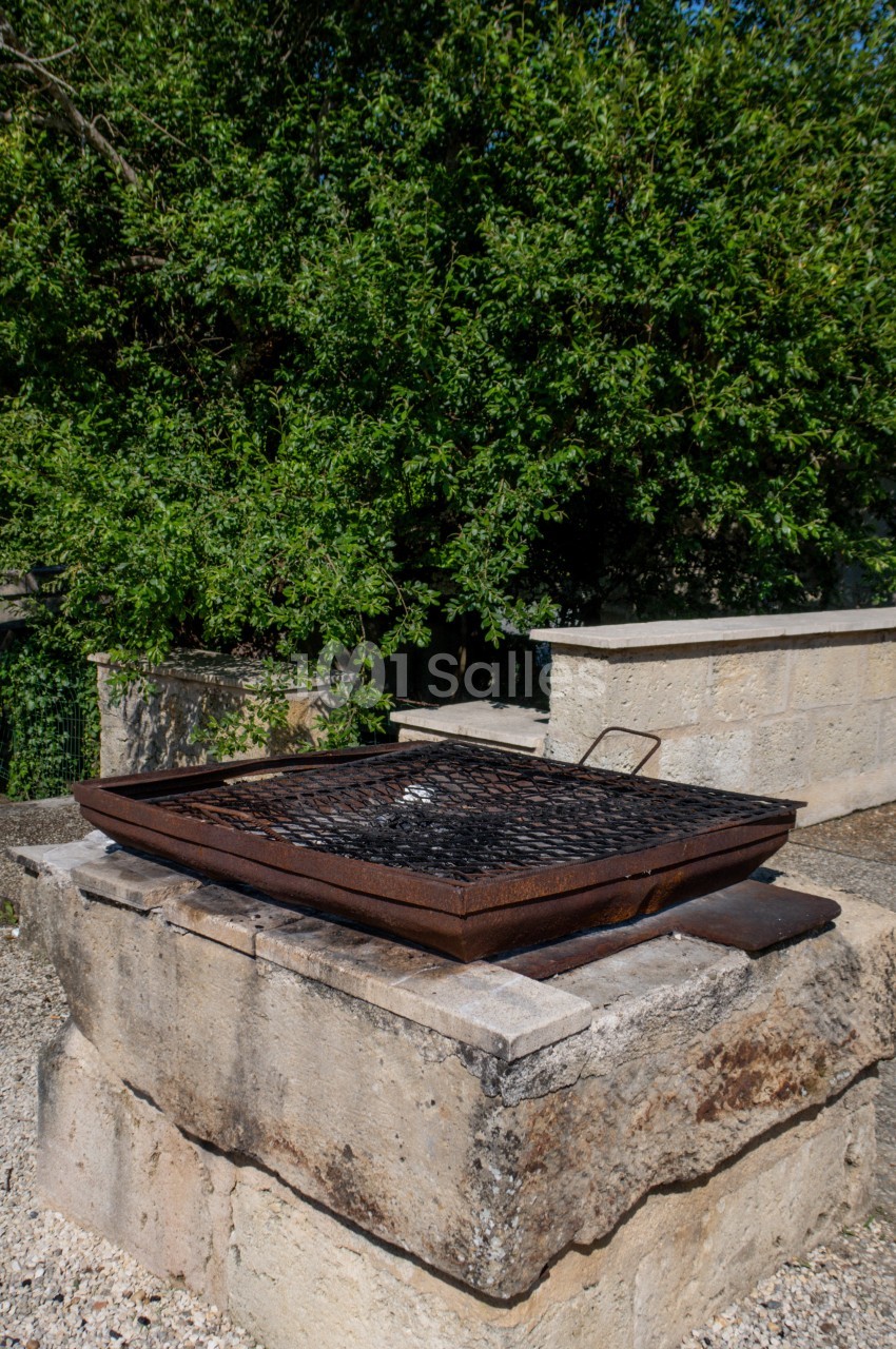 Barbecue en métal rouillé posé sur un socle en pierre, entouré de végétation et de murs en pierre.
