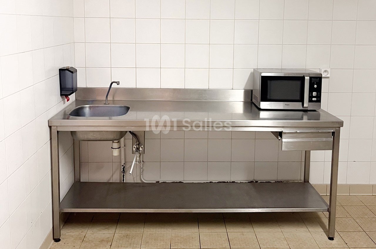 Évier en inox avec robinet, micro-ondes et plan de travail dans une cuisine aux murs carrelés blancs.