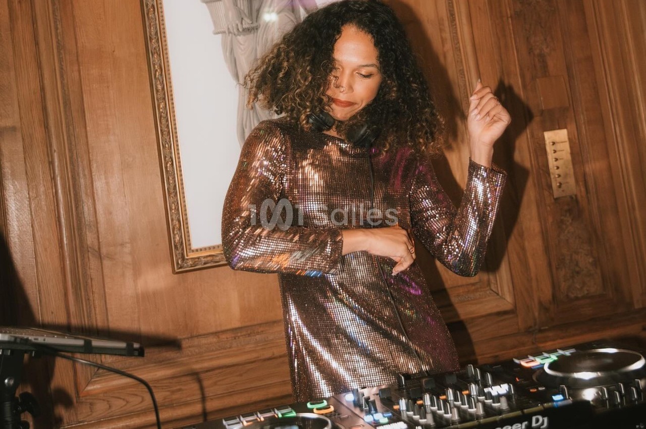 Une femme en robe scintillante mixe de la musique sur une table de DJ dans une pièce aux murs en bois.