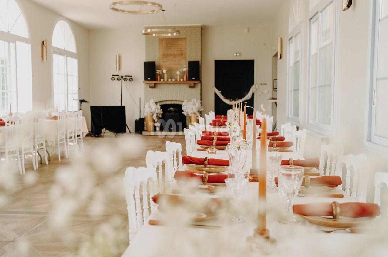 Salle de réception lumineuse avec de longues tables décorées de bougies, serviettes rouges et vaisselle élégante.