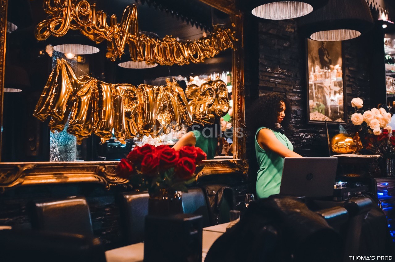 Femme en robe verte debout près d'un ordinateur, devant des ballons dorés et un miroir dans un décor de fête.