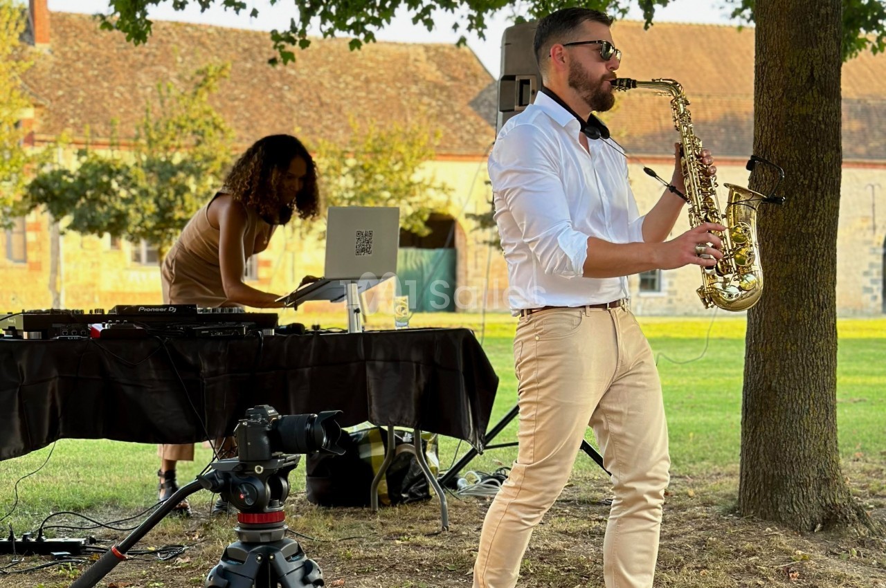Un musicien joue du saxophone en plein air tandis qu'une DJ mixe sur une table équipée d'un ordinateur portable.