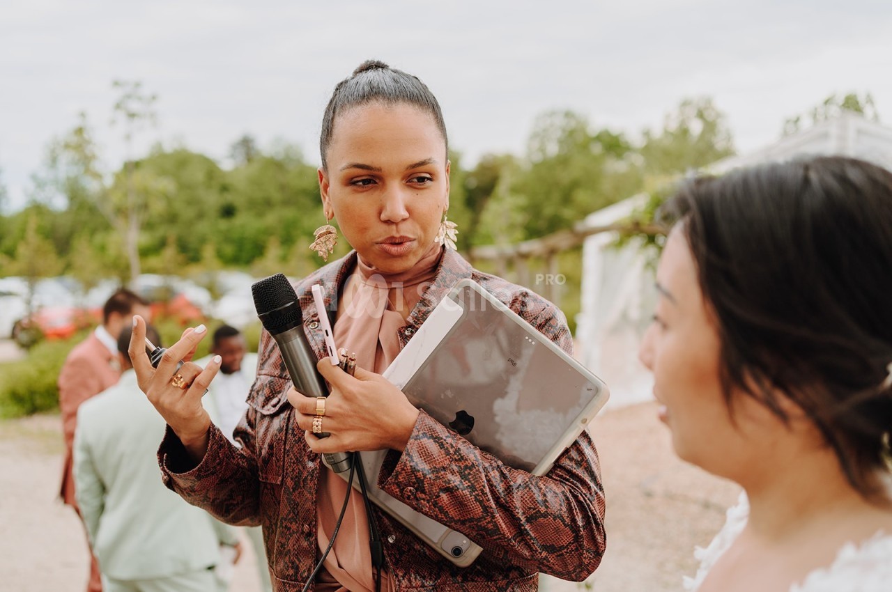 Une femme tenant un micro et une tablette parle à une autre personne en extérieur, avec des arbres en arrière-plan.