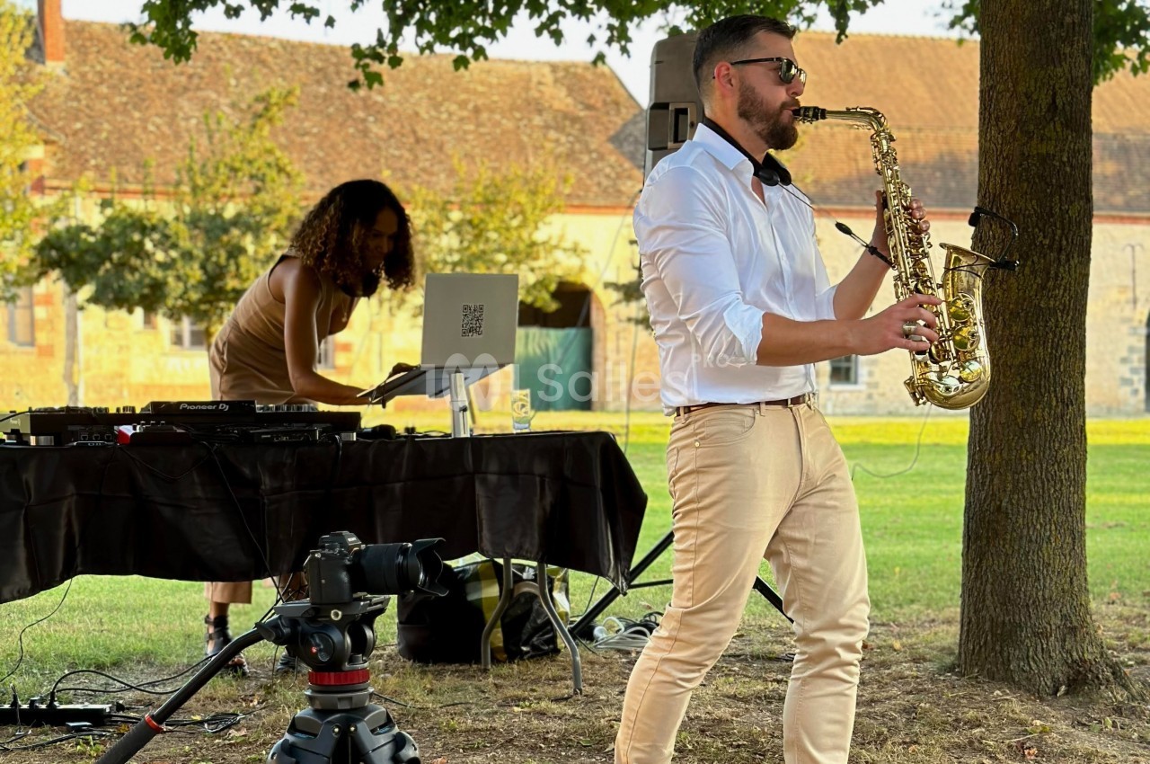 Un saxophoniste joue en plein air près d'une DJ animant une table de mixage, dans un cadre verdoyant.