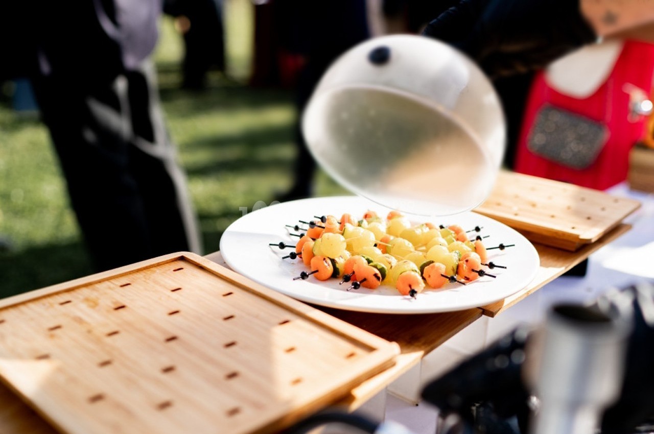 Assiette de brochettes de fruits colorés sous une cloche transparente, présentée sur une table en extérieur.
