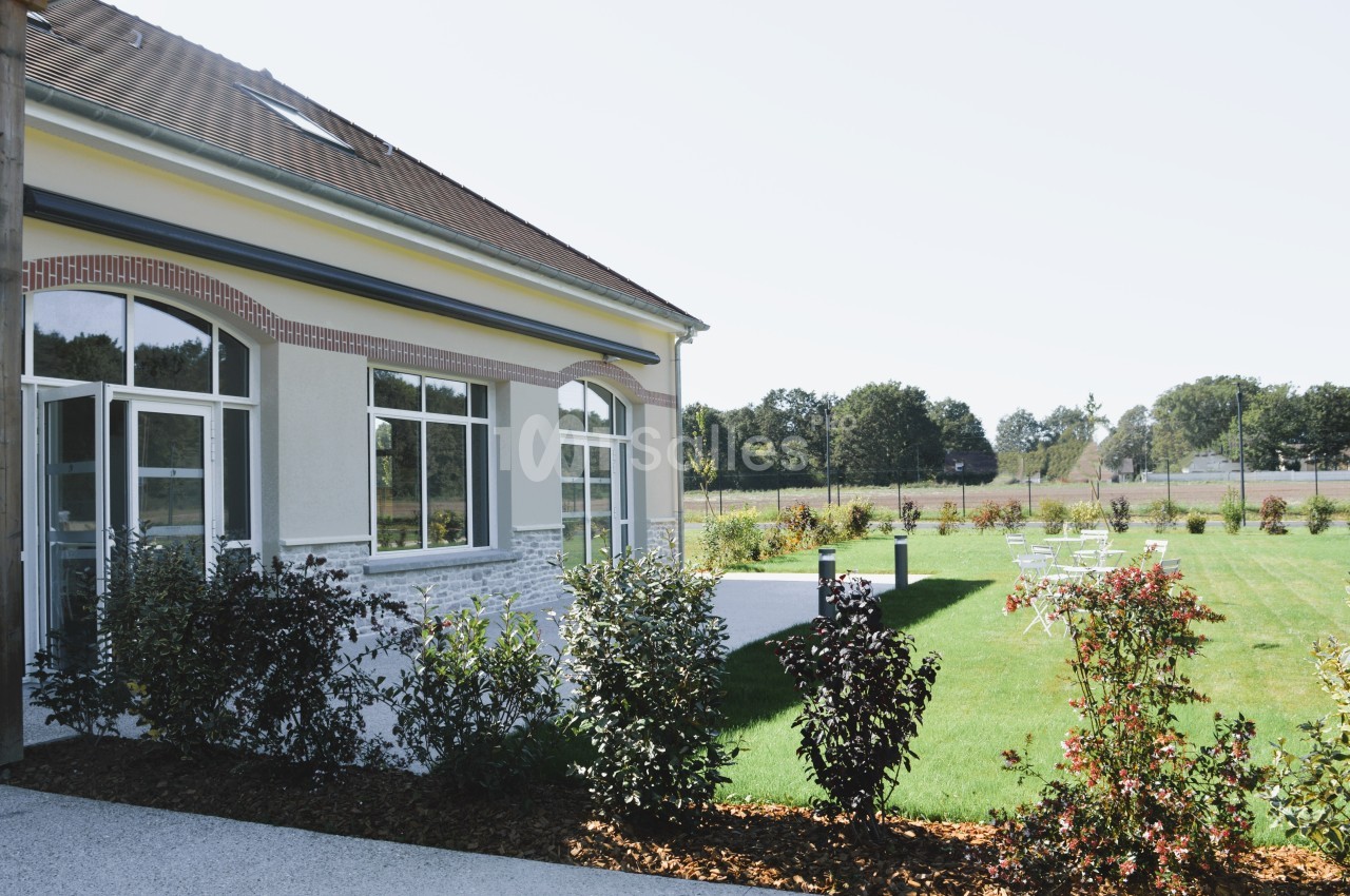 Bâtiment avec de grandes fenêtres donnant sur un jardin aménagé avec pelouse, arbustes et quelques chaises blanches.