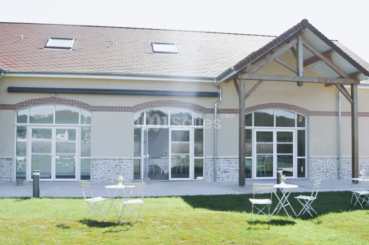 Façade d'un bâtiment moderne avec de grandes baies vitrées, terrasse aménagée avec tables et chaises sur pelouse.