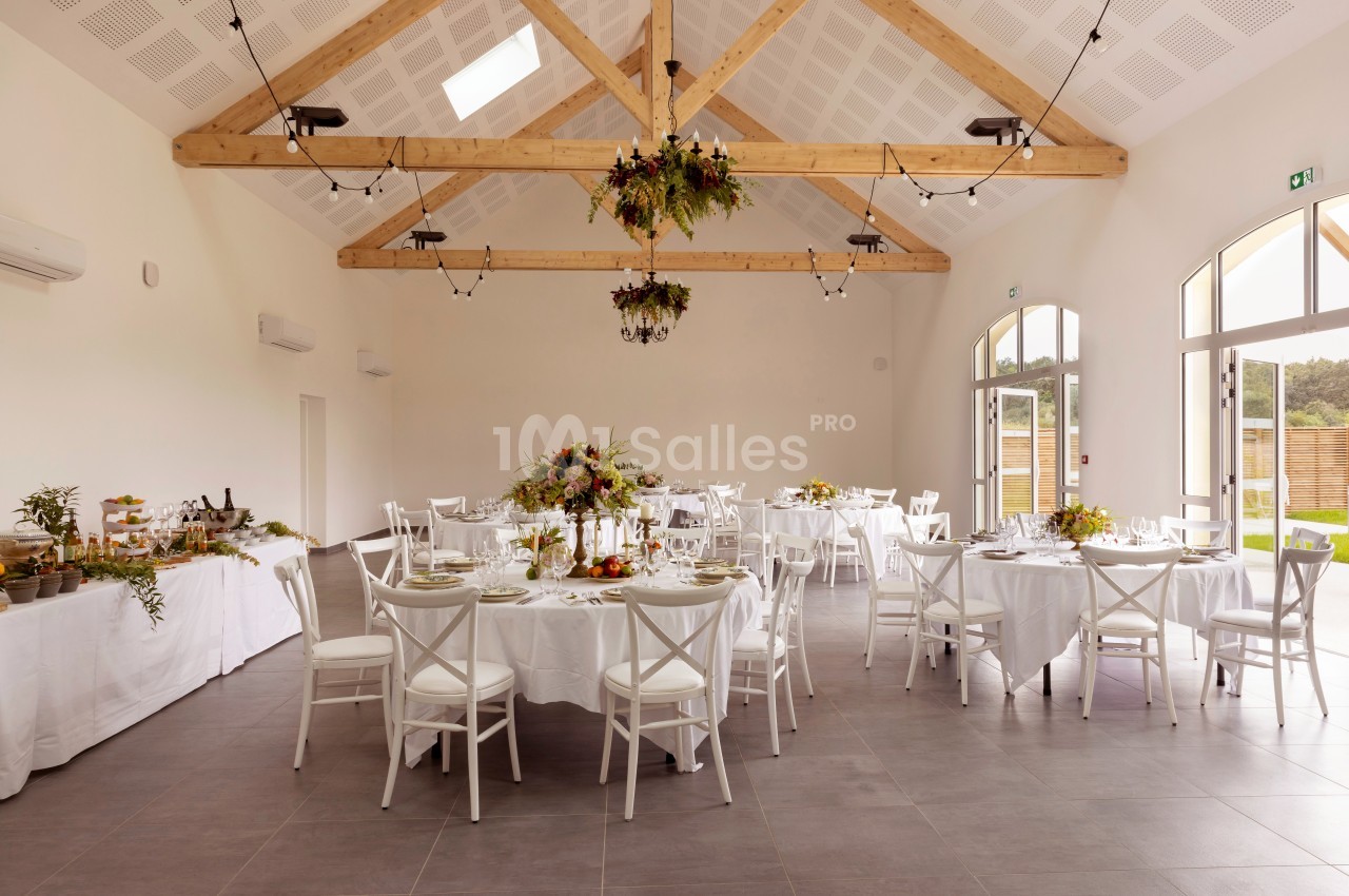 Salle lumineuse décorée pour un événement, avec tables rondes dressées et ornées de fleurs sous une charpente en bois.
