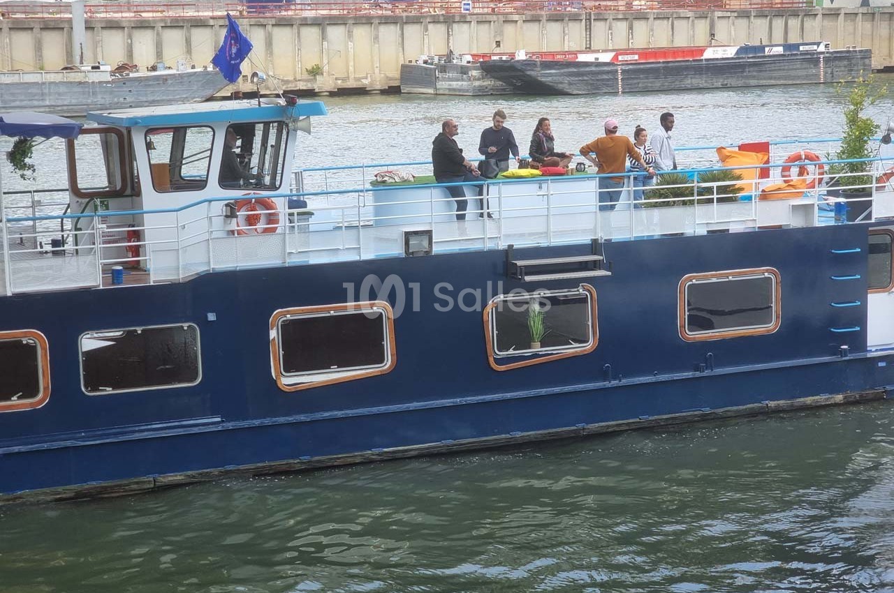 Bateau amarré sur un canal avec des personnes discutant sur le pont supérieur près de tables et de chaises.