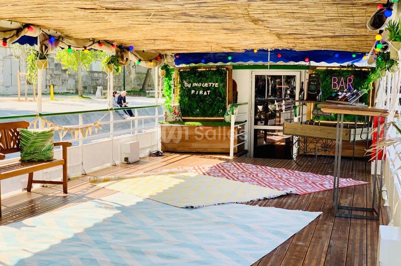 Terrasse en bois ombragée avec tapis colorés, banc en bois, décorations lumineuses et entrée d'un bar en arrière-plan.