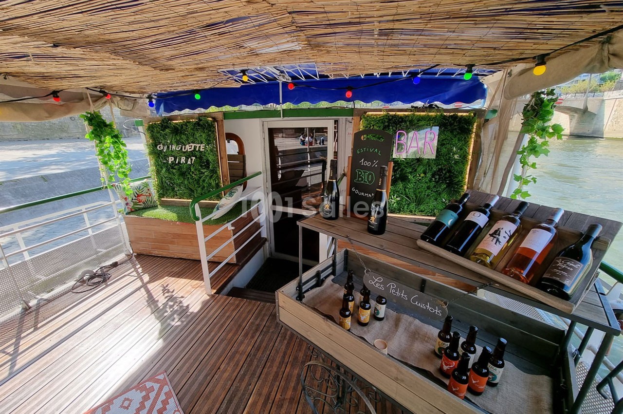 Terrasse d'une péniche aménagée avec un bar, des étagères de bouteilles de vin et une décoration végétale.