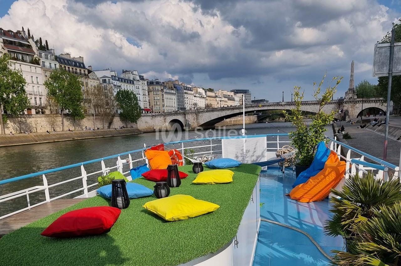 Terrasse colorée avec coussins et plantes sur une péniche, vue sur la Seine et des immeubles parisiens sous un ciel nuageux.