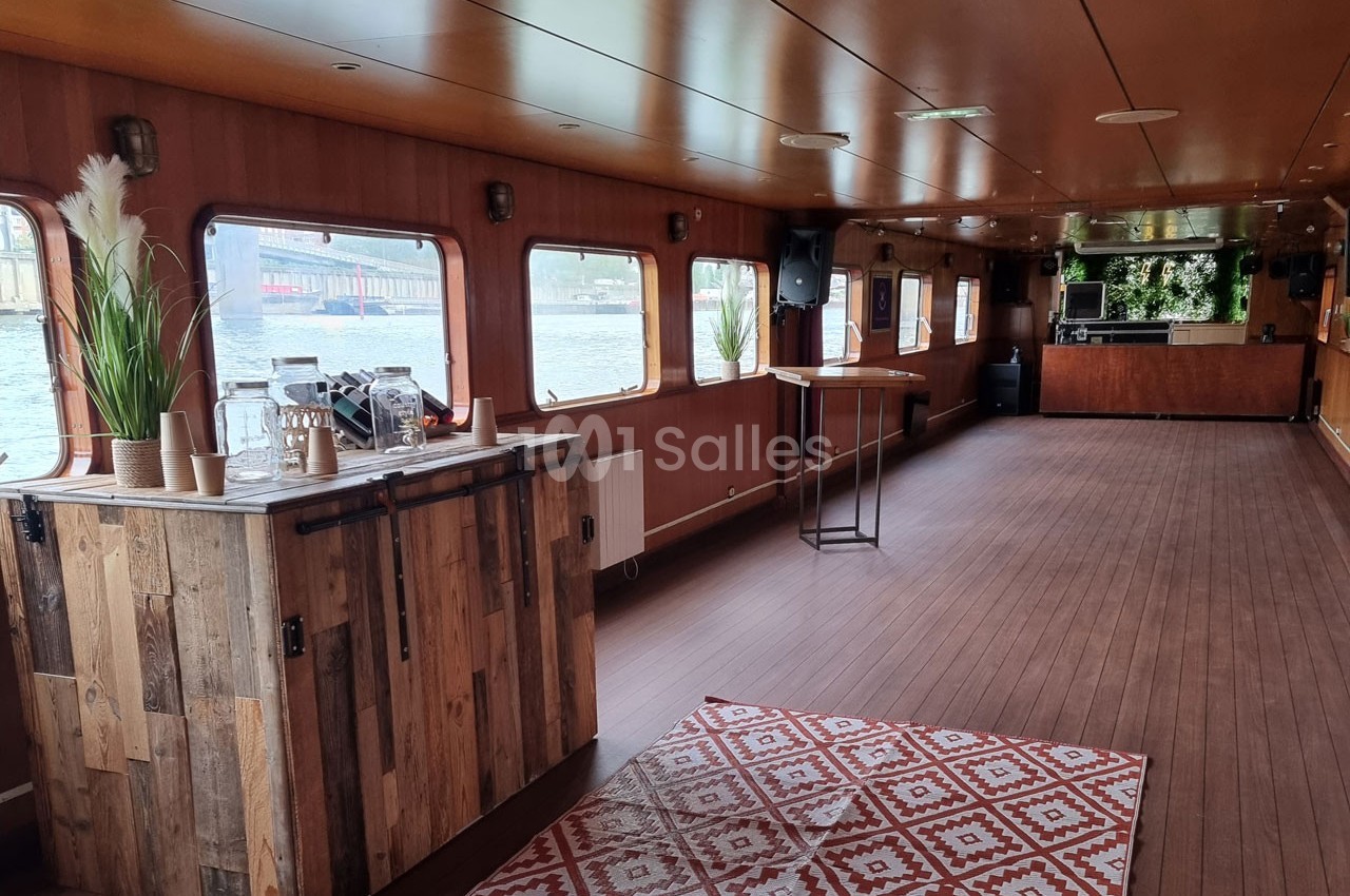 Intérieur d'une péniche avec parquet, bar en bois, tapis décoratif et grandes fenêtres donnant sur l'eau.