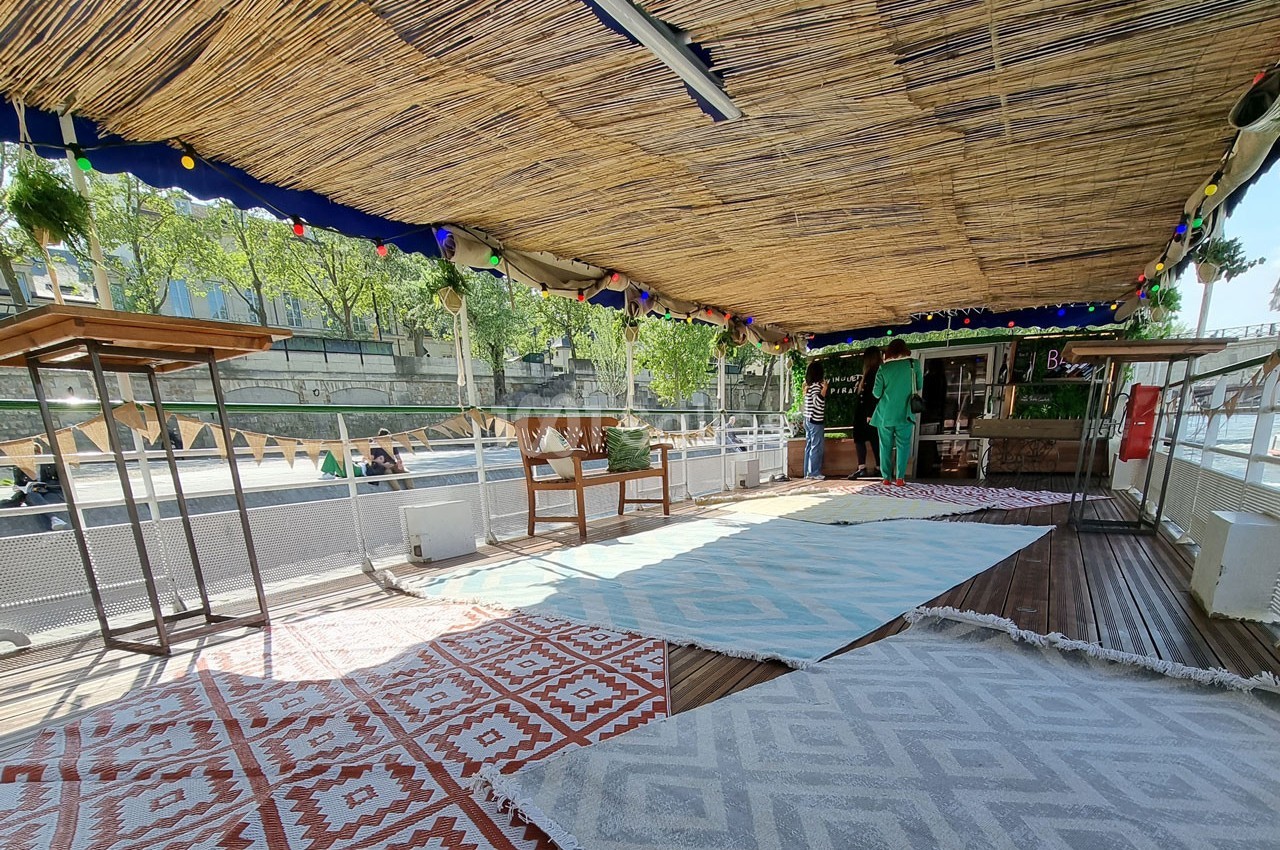 Terrasse couverte en bord de Seine avec tapis colorés, mobilier en bois et vue sur les quais et la végétation.