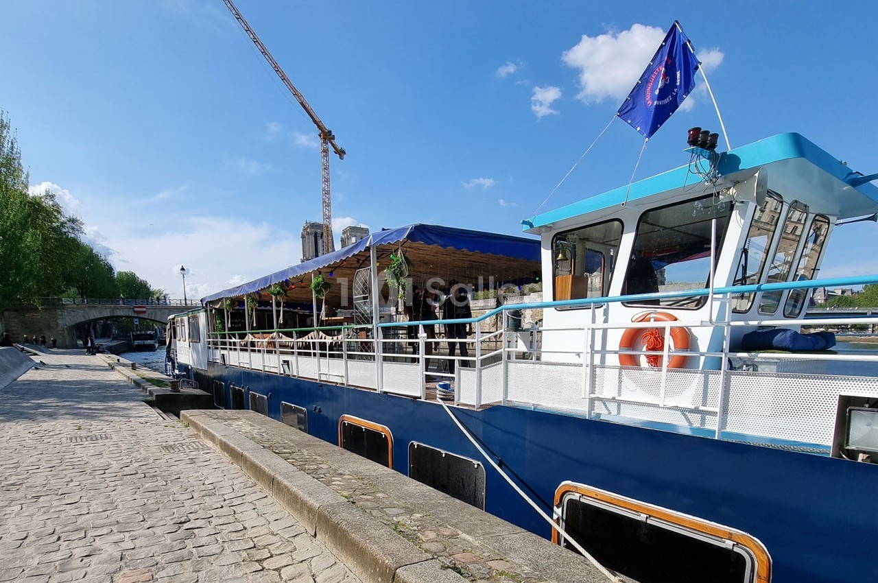 Bateau amarré le long d'un quai pavé, avec une terrasse couverte et une grue visible en arrière-plan.
