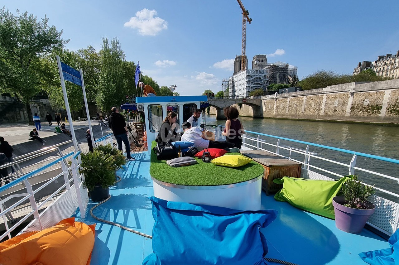 Des personnes assises sur un bateau coloré avec des coussins, naviguant sur une rivière bordée d'arbres et de bâtiments.