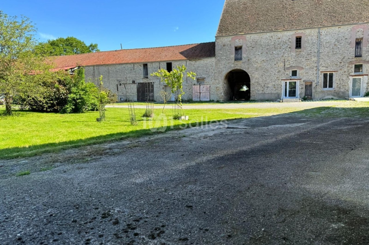 Cour d'une ferme ancienne avec bâtiments en pierre, pelouse, arbres et ciel bleu en arrière-plan.