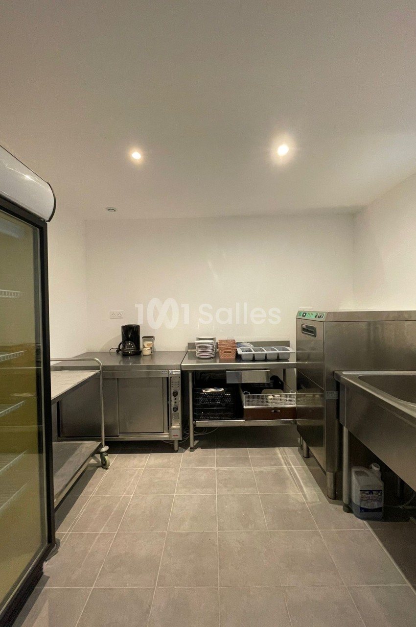 Cuisine professionnelle avec équipements en inox, évier, réfrigérateur vitré et plans de travail sous un éclairage lumineux.