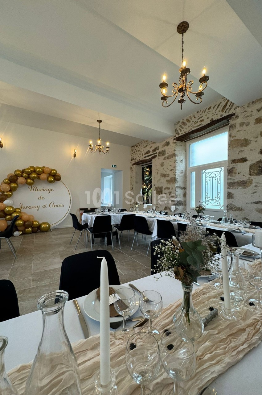 Salle de réception décorée pour un mariage, avec tables dressées, bougies, fleurs et arche ornée de ballons dorés.
